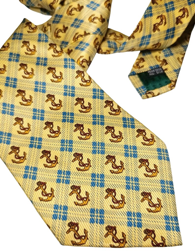 May include: A yellow silk tie with a repeating pattern of brown anchors and blue checkered squares. The tie is angled, showing the front and back, with a green tag visible.