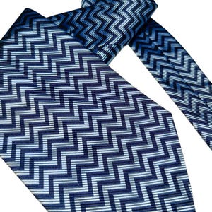 May include: A blue necktie featuring a repeating zigzag pattern in shades of blue and white. The tie is knotted, with the wider and narrower ends visible. The pattern gives a textured appearance.