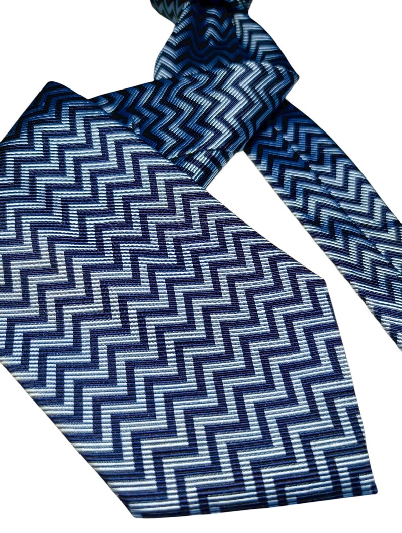 May include: A blue necktie featuring a repeating zigzag pattern in shades of blue and white. The tie is knotted, with the wider and narrower ends visible. The pattern gives a textured appearance.