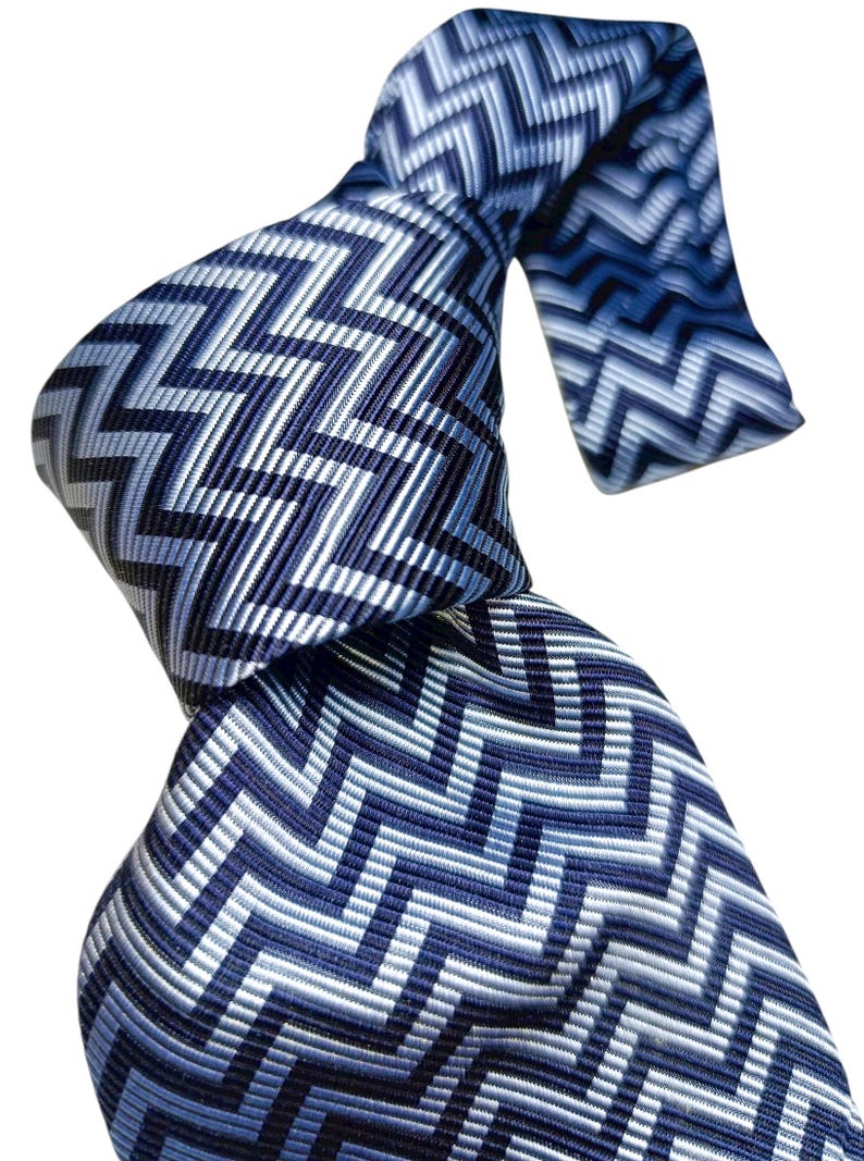 May include: A patterned necktie in shades of blue and white, featuring a zig-zag design. The tie is knotted, highlighting the intricate pattern and colour variations. The design incorporates various shades of blue and white, creating a stylish accessory.