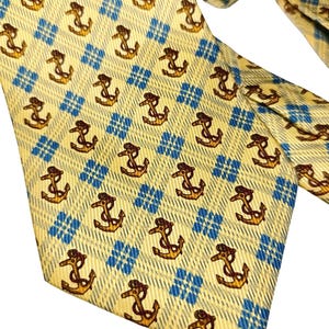 May include: A yellow tie with a repeating pattern of brown anchors and blue diamond shapes. The tie is made of a textured fabric and is knotted at the top, with the end of the tie visible.
