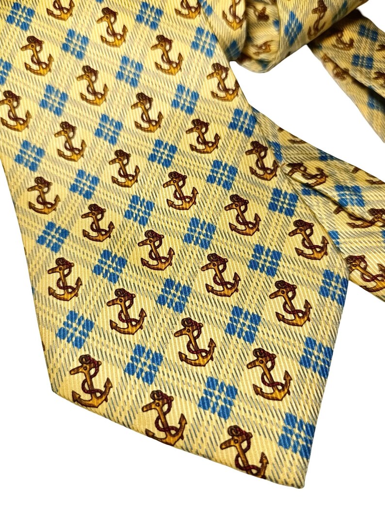 May include: A yellow tie with a repeating pattern of brown anchors and blue diamond shapes. The tie is made of a textured fabric and is knotted at the top, with the end of the tie visible.