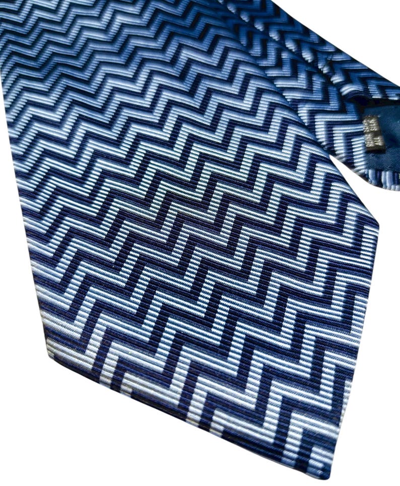 May include: A blue and white zigzag patterned tie. The tie displays a repeating chevron design in shades of blue and white. The fabric has a subtle sheen, and the tie is angled, showcasing the pattern's direction.