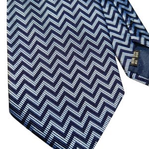 May include: A navy blue silk tie featuring a repeating chevron pattern in shades of blue and white. The tie is angled, displaying the intricate design and texture. The tie is made of silk.