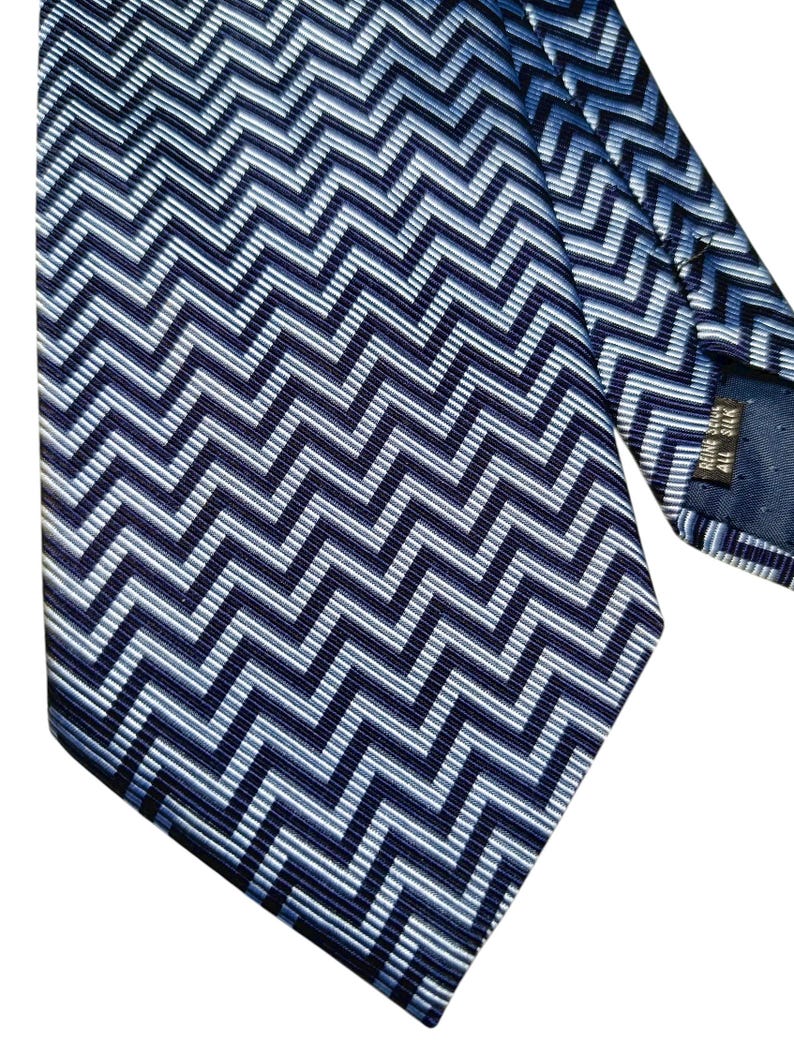 May include: A navy blue silk tie featuring a repeating chevron pattern in shades of blue and white. The tie is angled, displaying the intricate design and texture. The tie is made of silk.