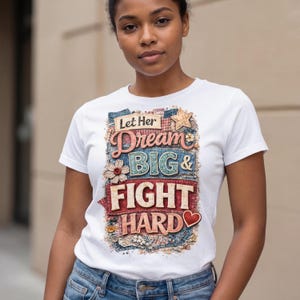 Puede incluir: Camiseta blanca con el texto "Let Her Dream BIG & FIGHT HARD" en un diseño de patchwork colorido. El gráfico incluye detalles florales y de estrella, y un corazón. La camiseta es de estilo cuello redondo clásico.