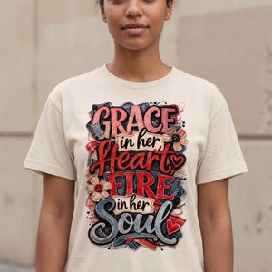 Puede incluir: Camiseta beige con el texto "GRACE in her HEART FIRE in her SOUL" en un diseño decorativo. El diseño incorpora elementos florales y de mezclilla en tonos rojos, rosas y azules. La camiseta está hecha de un material suave y cómodo.