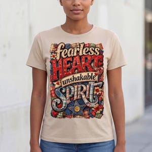 Puede incluir: Camiseta beige con las palabras "fearless HEART unshakable SPIRIT" en un diseño de patchwork. El diseño incluye detalles en rojo, azul y flores. La camiseta es de manga corta y tiene cuello redondo.