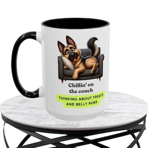 Może przedstawiać: Biały kubek ceramiczny z czarnym uchwytem i rantem. Kubek przedstawia kreskówkowego owczarka niemieckiego leżącego na kanapie z tekstem "Chillin' on the couch thinking about treats and belly rubs".