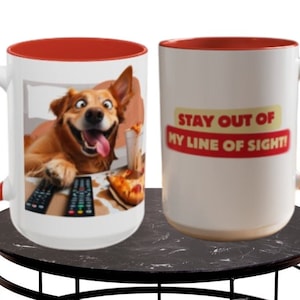 Może przedstawiać: Dwa białe ceramiczne kubki z czerwonymi obwódkami. Jeden kubek przedstawia psa z zabawnym wyrazem twarzy, trzymającego pilota i pizzę. Drugi kubek ma napis "STAY OUT OF MY LINE OF SIGHT!" w kolorze czerwonym i żółtym.