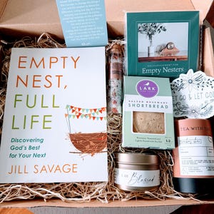 May include: A gift box containing a book titled Empty Nest, Full Life by Jill Savage, a Serenity Prayer card, a glass tube of bath salts, a box of Lark Salted Rosemary Shortbread, a candle labeled I Am Blessed, and a Tea With Tae container.