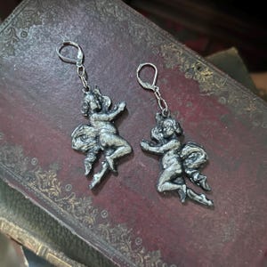 Peut inclure: Une paire de boucles d'oreilles argent&eacute;es repr&eacute;sentant des ch&eacute;rubins. Chaque boucle d'oreille repr&eacute;sente une petite figurine d&eacute;taill&eacute;e avec les bras tendus et un n&oelig;ud. Les boucles d'oreilles ont une fermeture &agrave; levier et sont pr&eacute;sent&eacute;es sur une surface sombre et textur&eacute;e.