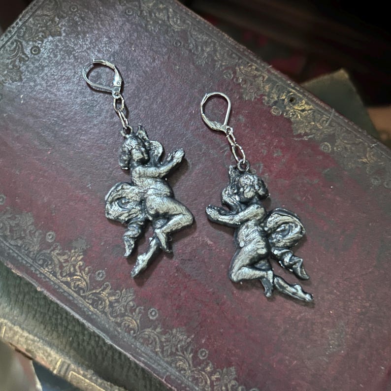 Peut inclure: Une paire de boucles d'oreilles argent&eacute;es repr&eacute;sentant des ch&eacute;rubins. Chaque boucle d'oreille repr&eacute;sente une petite figurine d&eacute;taill&eacute;e avec les bras tendus et un n&oelig;ud. Les boucles d'oreilles ont une fermeture &agrave; levier et sont pr&eacute;sent&eacute;es sur une surface sombre et textur&eacute;e.