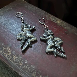 Peut inclure: Une paire de boucles d'oreilles argent&eacute;es repr&eacute;sentant des ch&eacute;rubins. Chaque boucle d'oreille repr&eacute;sente une petite figure ail&eacute;e dans une pose dynamique, suspendue &agrave; un simple crochet. Les boucles d'oreilles sont pr&eacute;sent&eacute;es sur une surface sombre et textur&eacute;e.