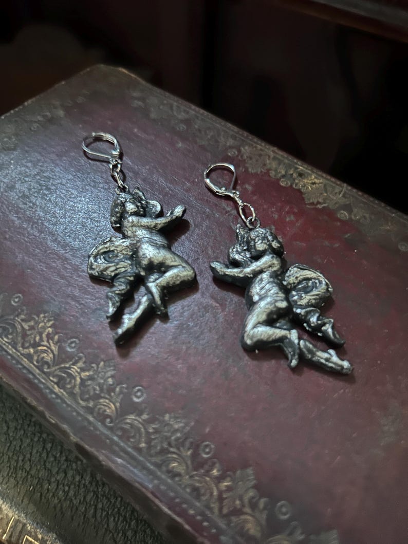 Peut inclure: Une paire de boucles d'oreilles argent&eacute;es repr&eacute;sentant des ch&eacute;rubins. Chaque boucle d'oreille repr&eacute;sente une petite figure ail&eacute;e dans une pose dynamique, suspendue &agrave; un simple crochet. Les boucles d'oreilles sont pr&eacute;sent&eacute;es sur une surface sombre et textur&eacute;e.