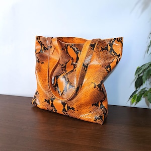 May include: A tote bag featuring a vibrant orange and black snakeskin pattern. The bag has two matching shoulder straps and a wide, open-top design. The textured surface mimics the appearance of reptile scales in a bold, high-contrast color scheme.