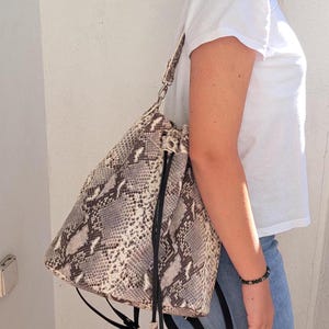 Puede incluir: Un bolso de cubo de piel de serpiente marrón y blanco con correas negras y un borla. El bolso se lleva sobre el hombro de una persona que lleva una camiseta blanca y unos vaqueros azules.
