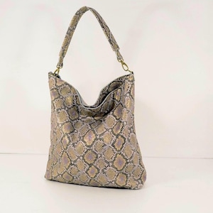 Może przedstawiać: Torba na ramię ze wzorem skóry węża w odcieniach złota, fioletu i szarości. Torba ma pojedynczy pasek na ramię i złote okucia. Torba wykonana jest z miękkiego, elastycznego materiału.