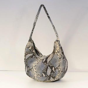 May include: A shoulder bag with a snakeskin pattern in shades of gray, white, and blue. The bag has a curved shape with a long strap and a zipper closure. The bag is made of a textured material.
