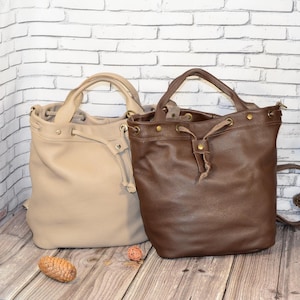 Puede incluir: Dos bolsos tipo cubo de cuero. Uno es de color beige claro y el otro marrón oscuro. Ambos bolsos tienen cierre de cordón, asas superiores y herrajes metálicos. Los bolsos están sobre una superficie de madera frente a una pared de ladrillos blancos.