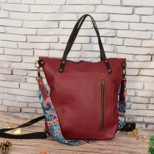 May include: A deep red textured tote bag featuring two black leather shoulder straps and a vertical zippered pocket on the front. The bag includes a detachable, wide shoulder strap with a colourful blue, orange, and teal paisley pattern.