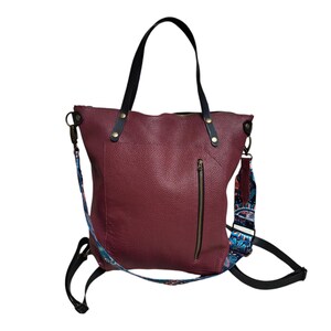 Peut inclure: Un sac cabas en cuir bordeaux avec une poignée en cuir noir et une bandoulière à motif coloré. Le sac a une poche zippée sur le devant.