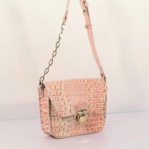 Puede incluir: Un bolso rosa con un estampado de piel de cocodrilo. El bolso tiene un cierre dorado y una correa de cadena. La correa para el hombro también es rosa y coincide con el color del bolso. El bolso cuelga sobre un fondo blanco.