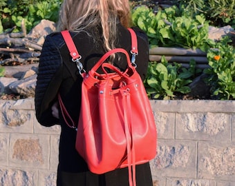 May include: A red leather backpack with a drawstring closure and adjustable straps. The bag has silver-colored hardware and is worn over the shoulders. The person is wearing a black coat.