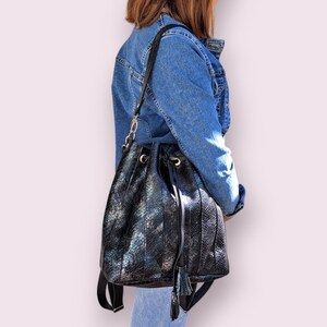 May include: Black bucket bag with a snakeskin pattern and a drawstring closure. The bag has a shoulder strap and a decorative tassel. The bag is made of a shiny, textured material.