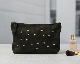 May include: A black suede cosmetic bag with a gold zipper and gold stud embellishments. The bag is rectangular and sits on a light-colored surface. A perfume bottle and makeup brush are in the background.