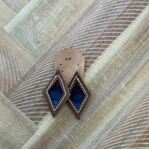 Pode incluir: Um par de brincos de couro castanho em forma de diamante. Os brincos são decorados com um padrão tecido azul e vermelho e têm costuras brancas ao redor das bordas.
