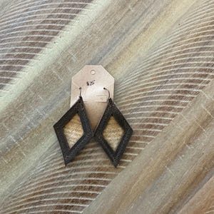 Pode incluir: Um par de brincos de couro marrom em forma de diamante com um centro de tecido bege. Os brincos estão pendurados em uma etiqueta de papel marrom com o número "#25" escrito nela.