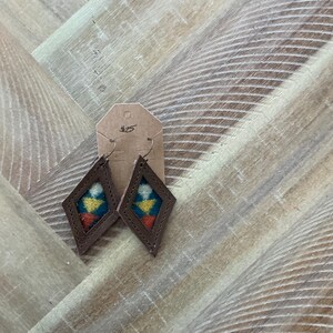 Pode incluir: Um par de brincos de couro castanho em forma de diamante. Os brincos têm um padrão tecido colorido no centro. A etiqueta de preço diz 25 €.