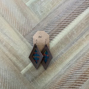 Pode incluir: Um par de brincos de couro marrom em forma de diamante com um padrão geométrico colorido no interior. Os brincos estão em uma etiqueta branca com o número 426 escrito nela.