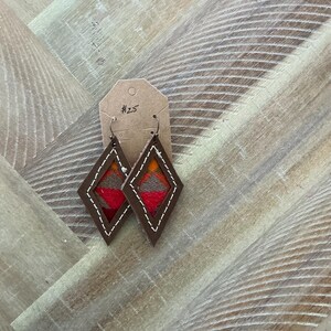 Pode incluir: Um par de brincos de couro marrom em forma de diamante. Os brincos são decorados com um inserto de tecido colorido com padrões. Os brincos são etiquetados com o número "#25".