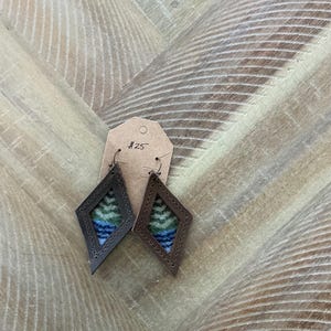 Pode incluir: Um par de brincos de couro marrom em forma de diamante com um padrão tecido azul, verde e branco no interior. Os brincos estão pendurados em uma etiqueta marrom com o número "#25" escrito nela.