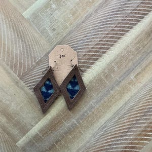 Pode incluir: Um par de brincos de couro castanho em forma de diamante. Os brincos têm um inserto de tecido com padrão azul e preto. A etiqueta de preço diz 25 €.