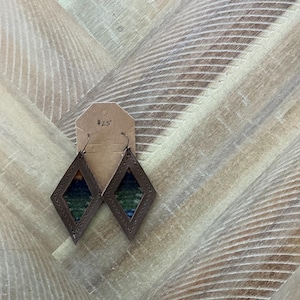 Pode incluir: Um par de brincos de couro marrom com formato de diamante e um inserto de tecido estampado colorido. Os brincos estão pendurados em um cartão marrom com a etiqueta de preço "2,5 €".