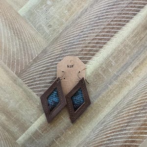 Pode incluir: Um par de brincos de couro marrom com formato de diamante. Os brincos têm um design estampado azul e marrom dentro da forma de diamante.