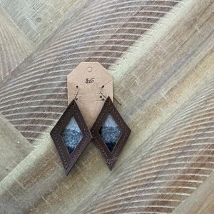 Pode incluir: Um par de brincos de couro marrom em forma de diamante. Os brincos são preenchidos com um tecido estampado em cinza e branco. Os brincos estão pendurados em uma etiqueta marrom com o preço "25 €" escrito nela.