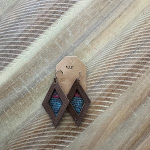 Pode incluir: Um par de brincos de couro marrom em forma de diamante com um inserto de tecido estampado em azul e vermelho. Os brincos têm um fecho de gancho prateado.