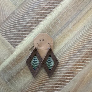 Pode incluir: Um par de brincos de couro castanho com forma de diamante. Os brincos têm um inserto de tecido com padrão verde e azul. A etiqueta de preço diz 25 €.