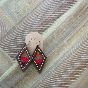 Pode incluir: Um par de brincos em forma de diamante com suporte de couro marrom e um inserto de tecido tecido colorido. Os brincos são etiquetados com o número "#25".