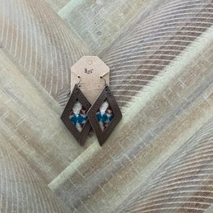 Pode incluir: Um par de brincos de couro marrom com formato de diamante. Os brincos apresentam um padrão tecido colorido dentro do couro.