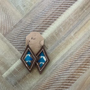Pode incluir: Um par de brincos de couro marrom em forma de diamante. Os brincos têm um inserto de tecido com padrão azul e branco. A etiqueta nos brincos diz "#25".