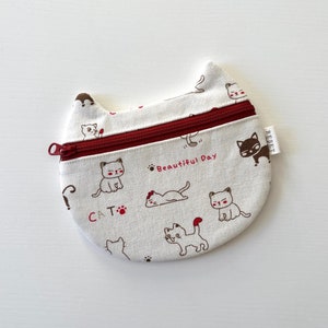 Puede incluir: Una cartera de monedas de tela blanca con forma de gato, con una cremallera roja y un estampado de gato marrón. La cartera tiene el texto "Beautiful Day" y "CAT" impreso.