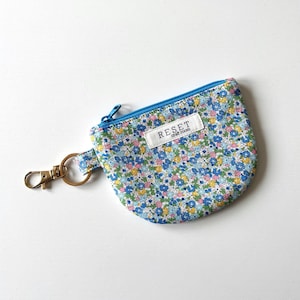 May include: A small, floral-patterned coin purse with a blue zipper and a gold-colored keychain attachment. The purse features a white label with the text "RESET craft room". The floral pattern includes blue, yellow, and pink flowers.