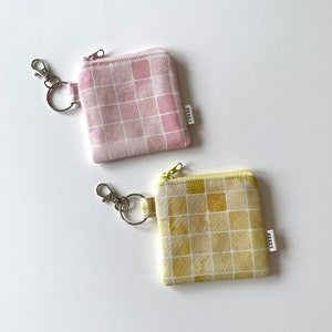 May include: Two small fabric pouches with keychains. Both pouches are square and have a checkered pattern. One pouch is pink with a white checkered pattern and the other is yellow with a white checkered pattern. The pouches have the word "REBET" printed on them.