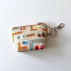 May include: A beige fabric coin purse with a zipper closure and a brown and white patterned design. The purse has a small key ring attached to it. The pattern includes a variety of small images, including a house, a flower, a bird, and a heart. The text "READY GO!!" is printed on the purse.