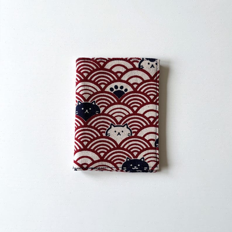 May include: A red and white fabric wallet with a pattern of overlapping circles and cats. The cats are black and white and have different expressions.